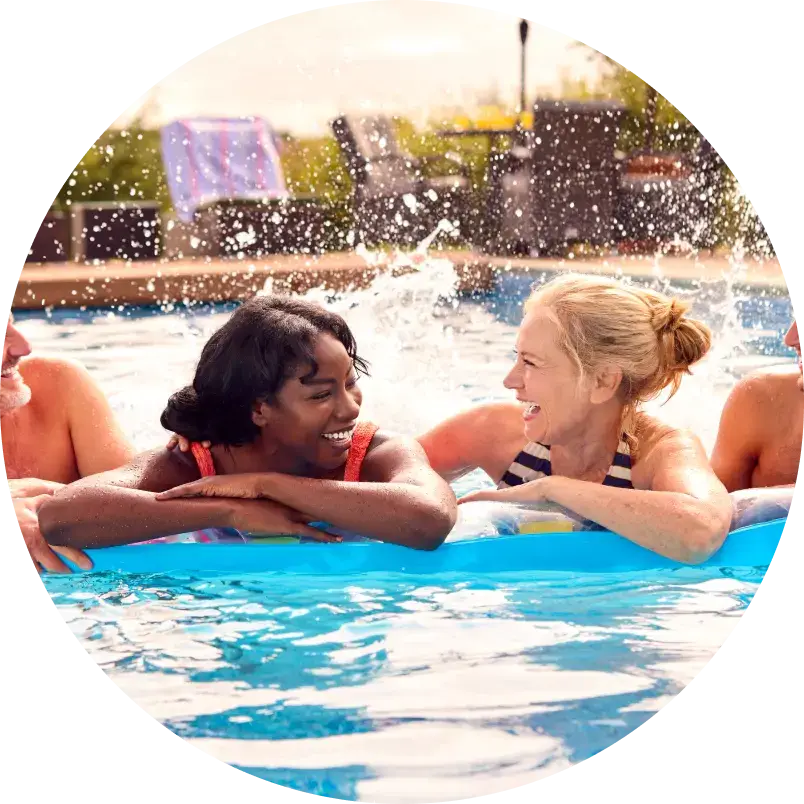 Two women smile and chat while leaning on pool floats in a sunlit pool. Water splashes around them, evoking a joyful and relaxed atmosphere.