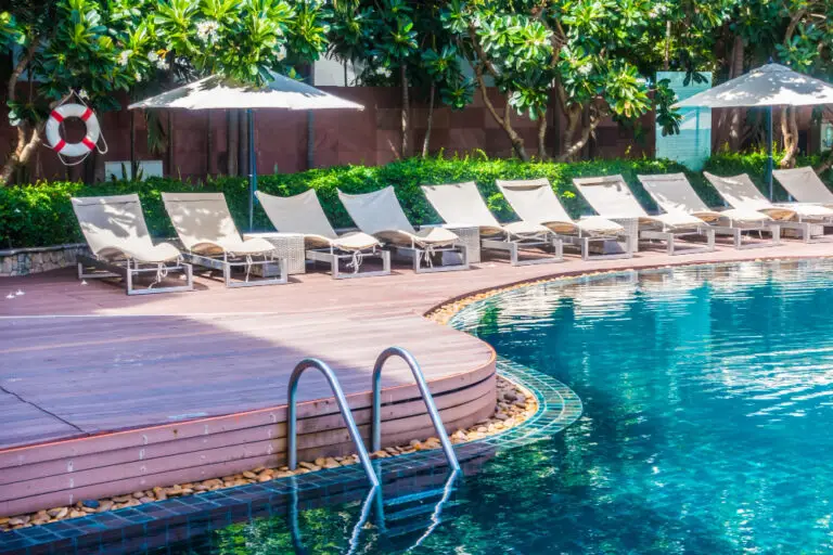 Sunny poolside scene with blue water, curved wooden deck, and beige lounge chairs. Umbrellas and lush greenery add a relaxing, inviting vibe.