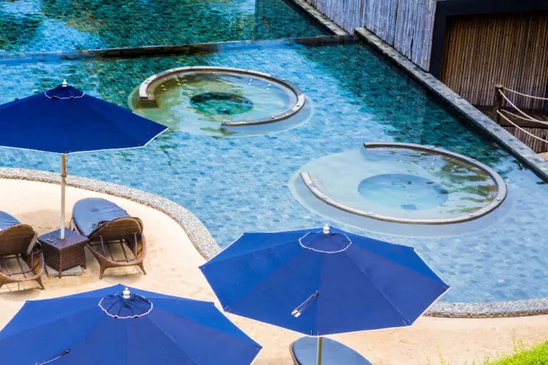 Blue umbrellas and cozy wicker chairs on a sandy patio overlook a serene pool with circular stone hot tubs, creating a tranquil, relaxing setting.
