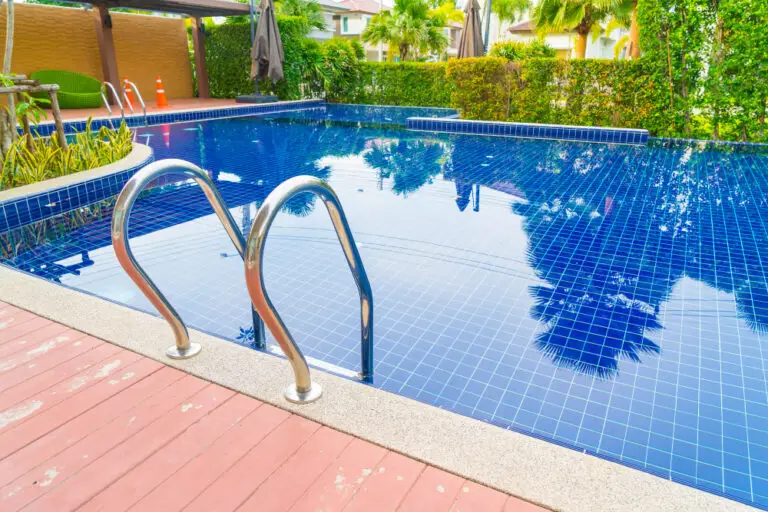 Sparkling outdoor pool with blue tiles and metal ladder, surrounded by lush greenery and a wooden deck, creates a tranquil and inviting atmosphere.