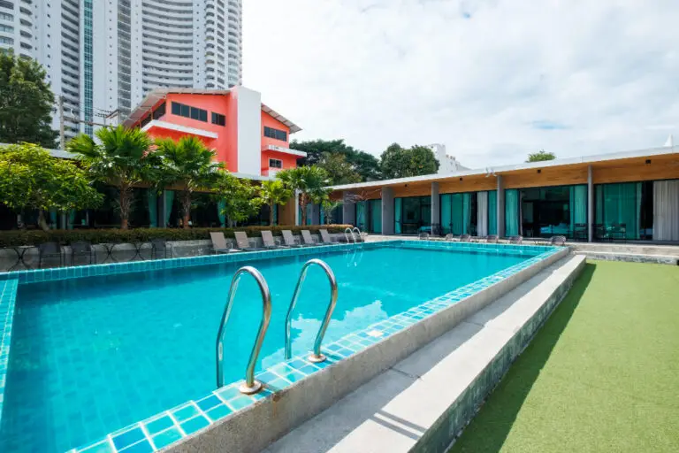 Outdoor pool with clear blue water adjacent to a modern building. Sun loungers line one side, with green trees and a tall white building in the background.