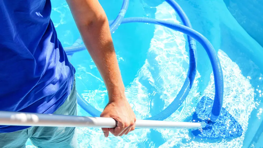 A person in a blue shirt cleans a bright blue swimming pool with a skimmer net.