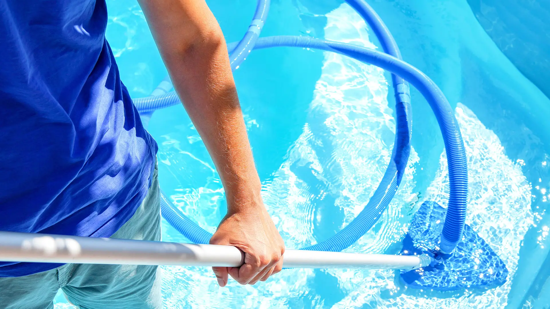 A person in a blue shirt cleans a bright blue swimming pool with a skimmer net.