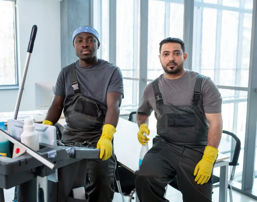 Two janitors wearing overalls and yellow gloves sit beside a cleaning cart with supplies in a bright office.