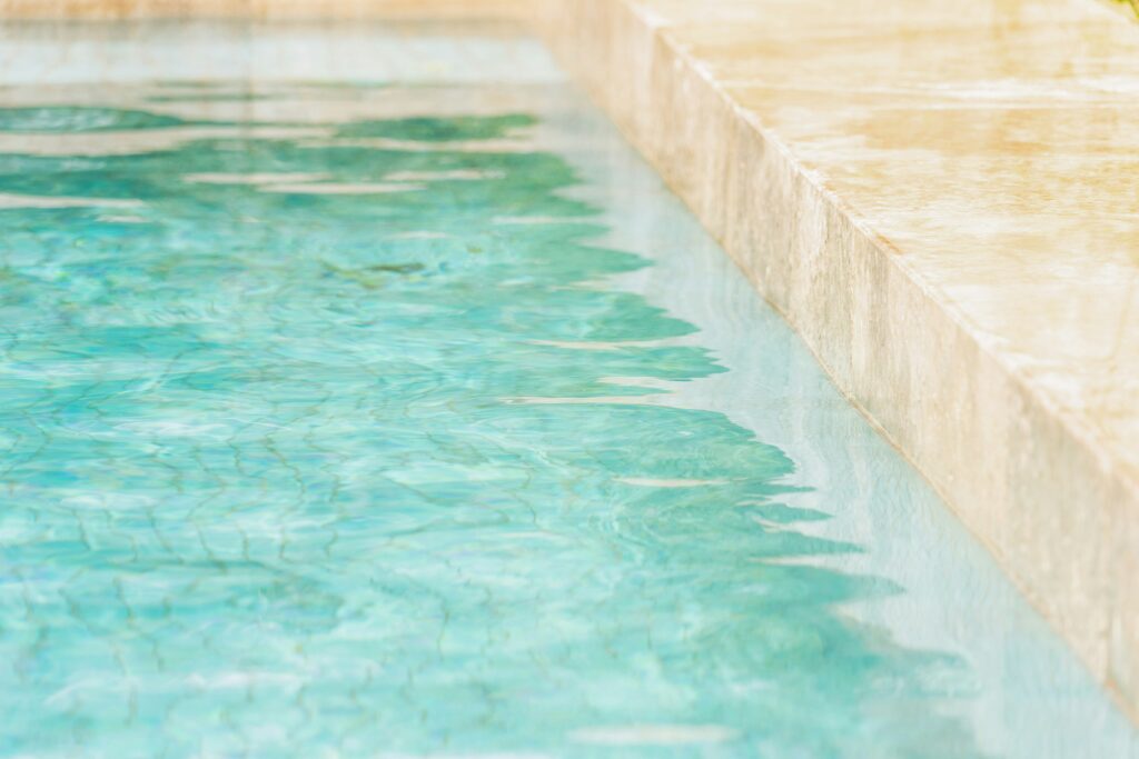 Clear turquoise pool water with gentle ripples, alongside a beige stone pool edge. The scene evokes a calm, refreshing, and inviting atmosphere.
