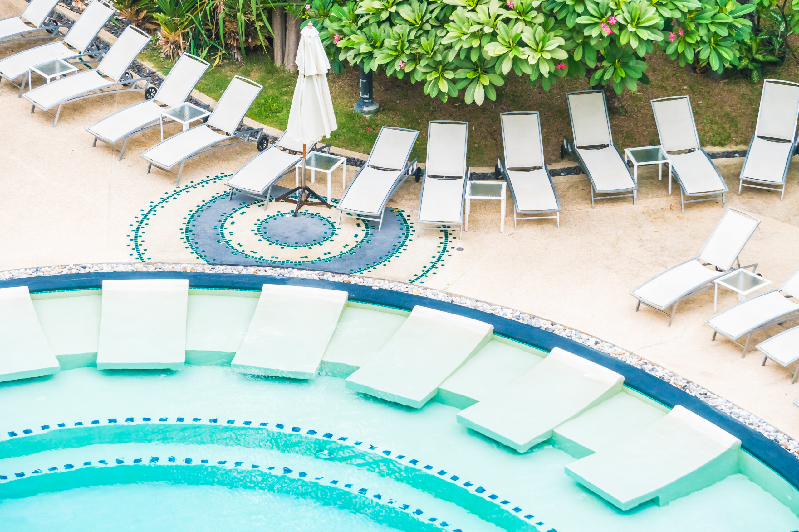 A circular pool with green water features reclined lounge chairs partially submerged. Surrounding the pool, more lounge chairs face lush green foliage. Serene ambiance.